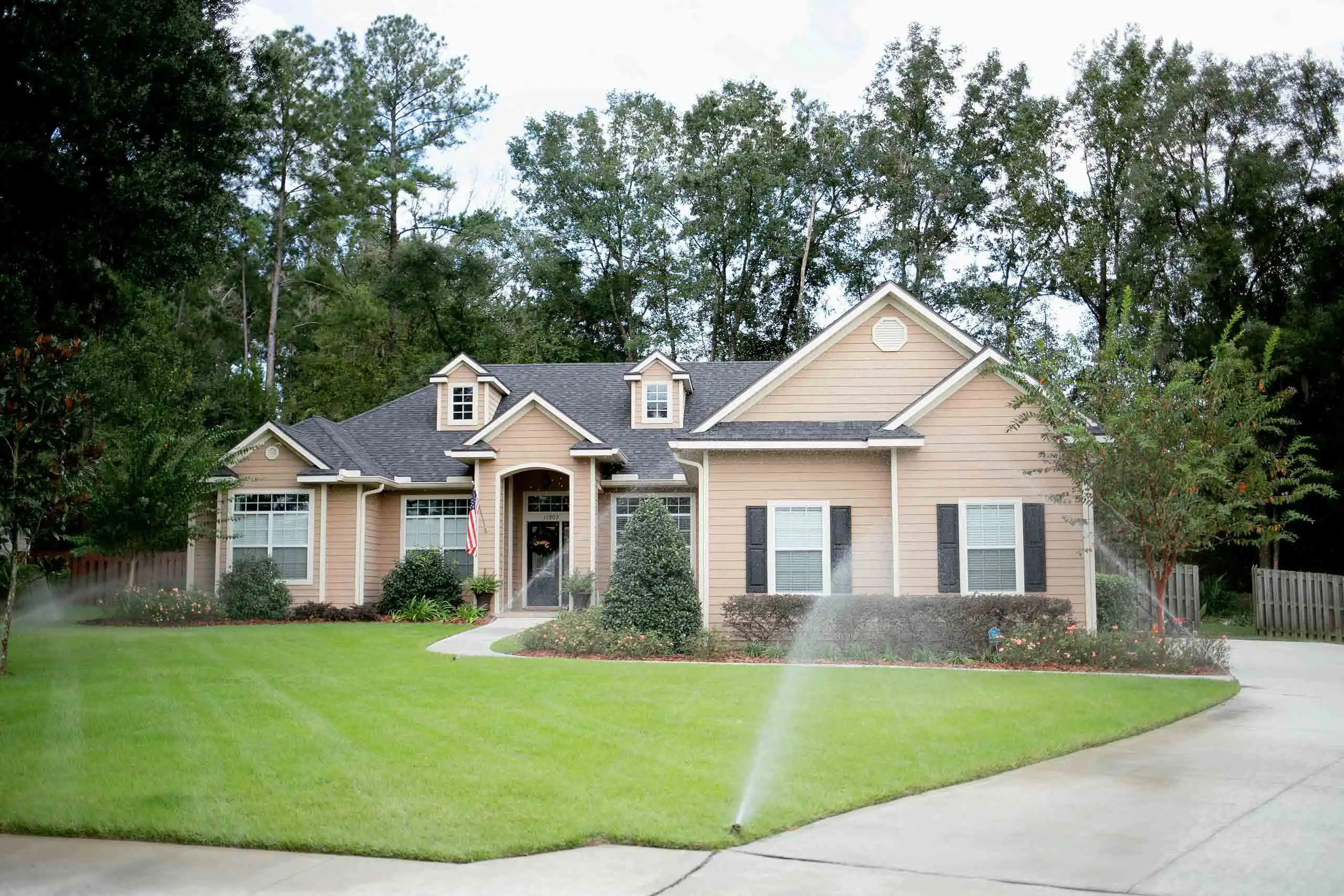 A single-story tan home with black shutters and a gabled roof, set behind a healthy green lawn with active sprinklers running and a curving driveway.