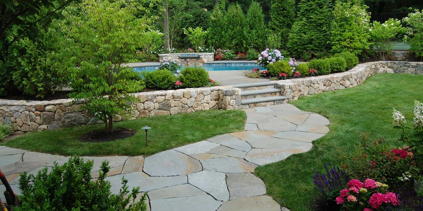 Beautiful backyard landscape with a stone walkway leading to a raised pool area, surrounded by stone retaining walls, lush green lawn, colorful flowers, and vibrant shrubs.