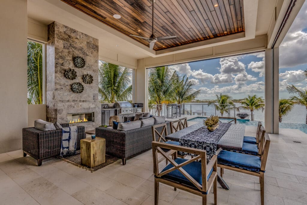 Luxury covered outdoor living space with wicker lounge seating, a modern fireplace, dining area, and built-in grill, overlooking a tropical waterfront with palm trees and an infinity pool.