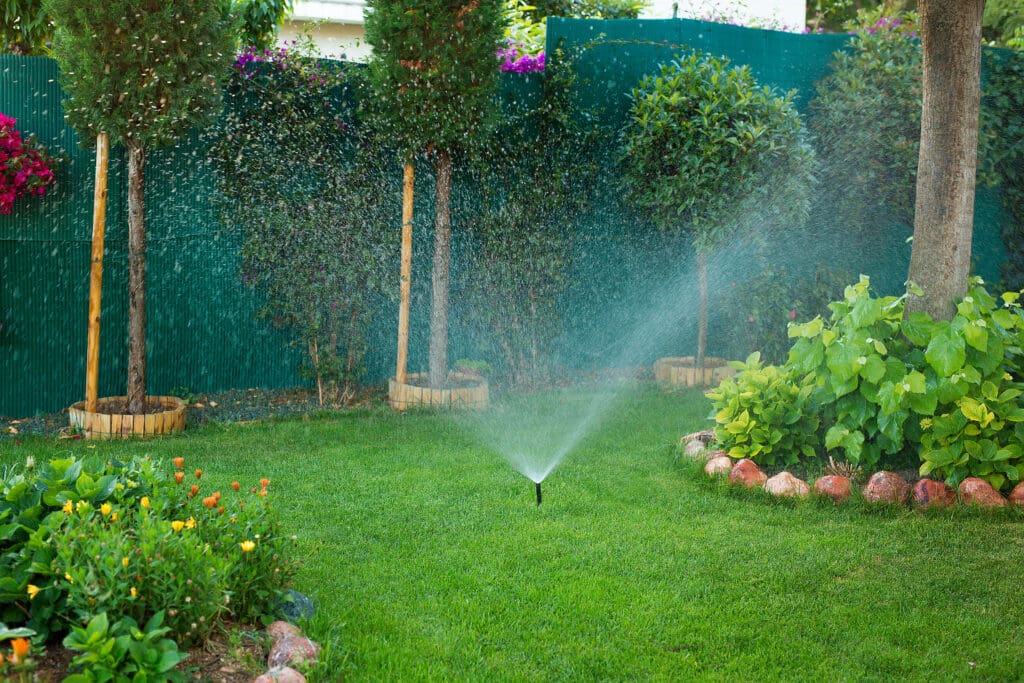 Sprinkler watering a lush green lawn bordered by flower beds and small trees, creating a vibrant backyard garden scene.