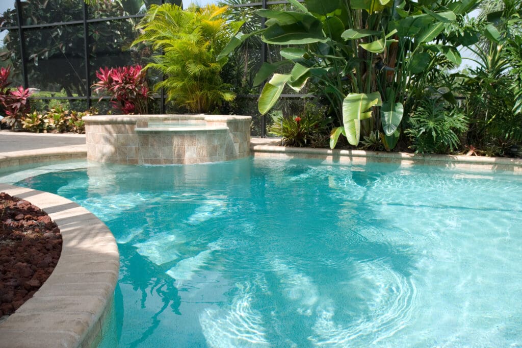 Tropical backyard pool surrounded by lush greenery and large leafy plants, with a built-in spa and serene blue water.