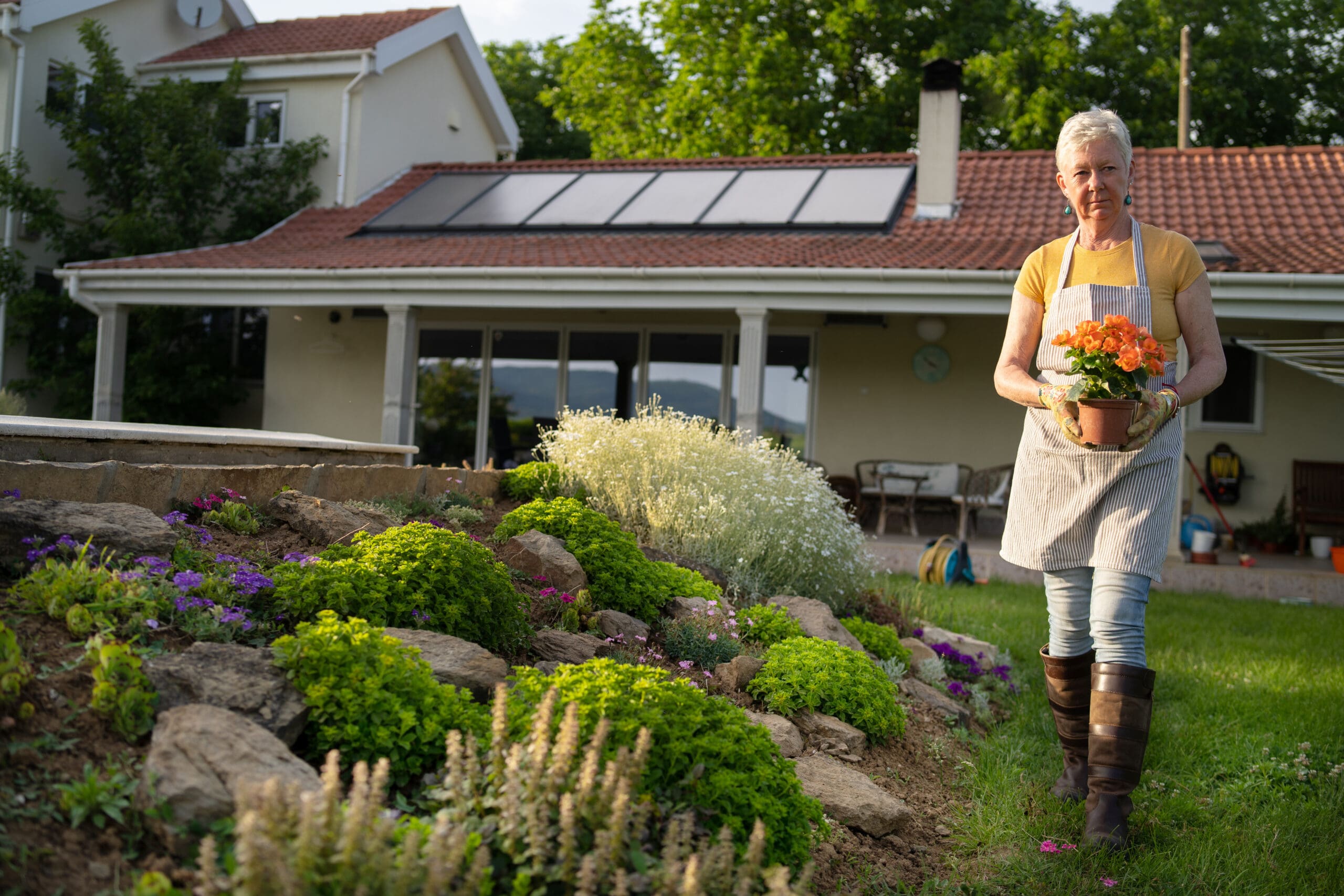 A woman in gardening attire holds a potted orange flower while walking through a vibrant garden filled with lush greenery and colorful blooms, with a house featuring a tiled roof and solar panels in the background.