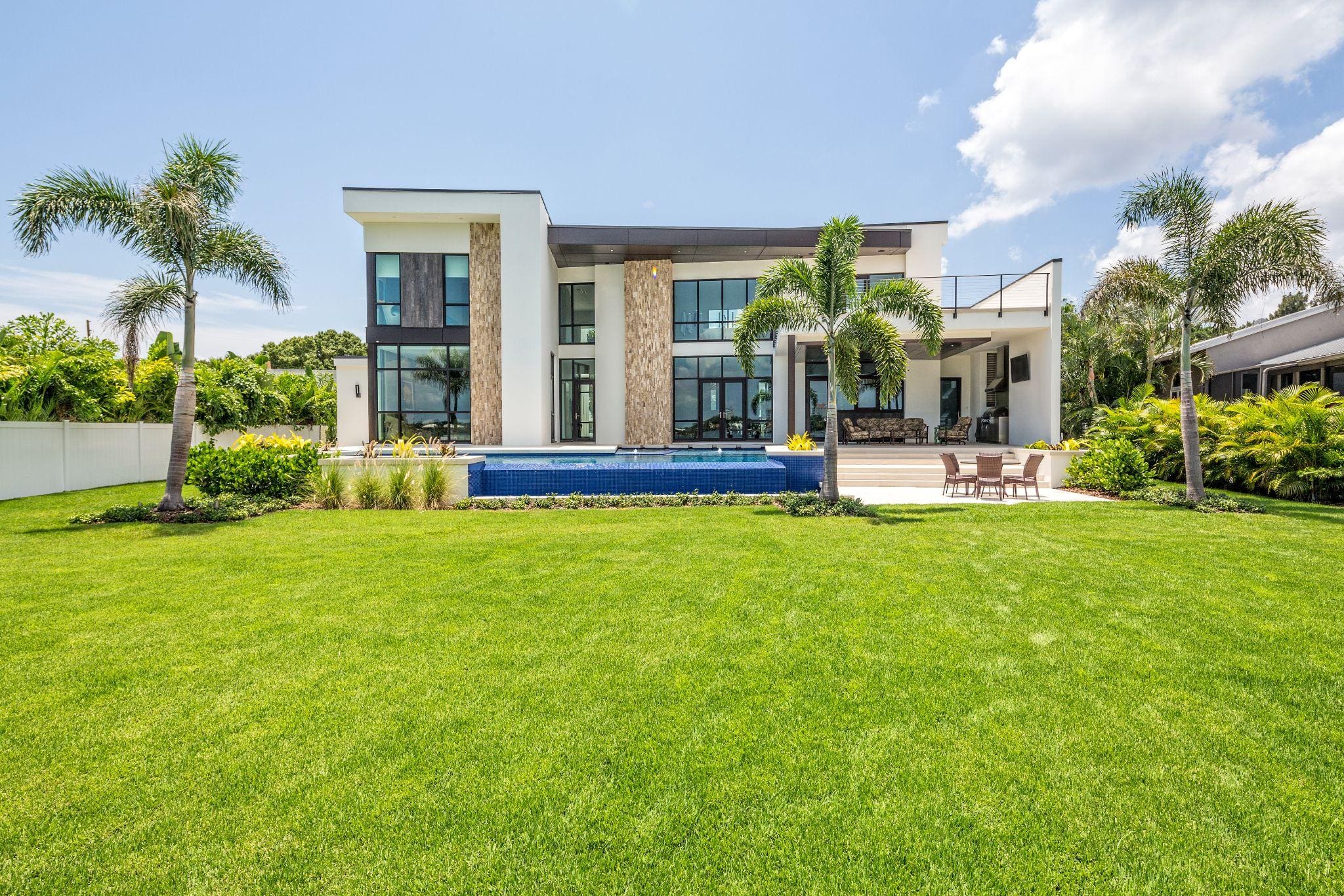 A modern two-story house with large windows and a sleek design, featuring a swimming pool in the backyard surrounded by lush green grass and palm trees under a clear blue sky.
