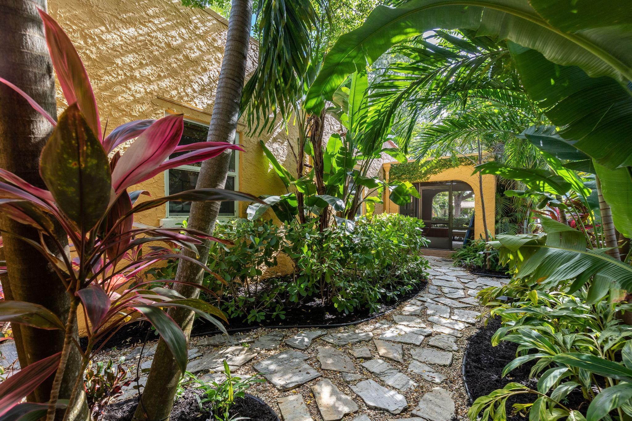 A pathway leading to a cozy entrance framed by tropical plants, featuring lush greenery and colorful foliage.