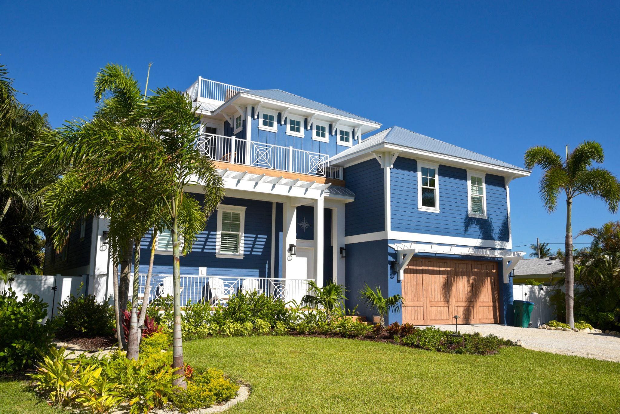 A stunning blue house with a wooden garage door and a balcony, framed by tropical plants and clear blue skies.