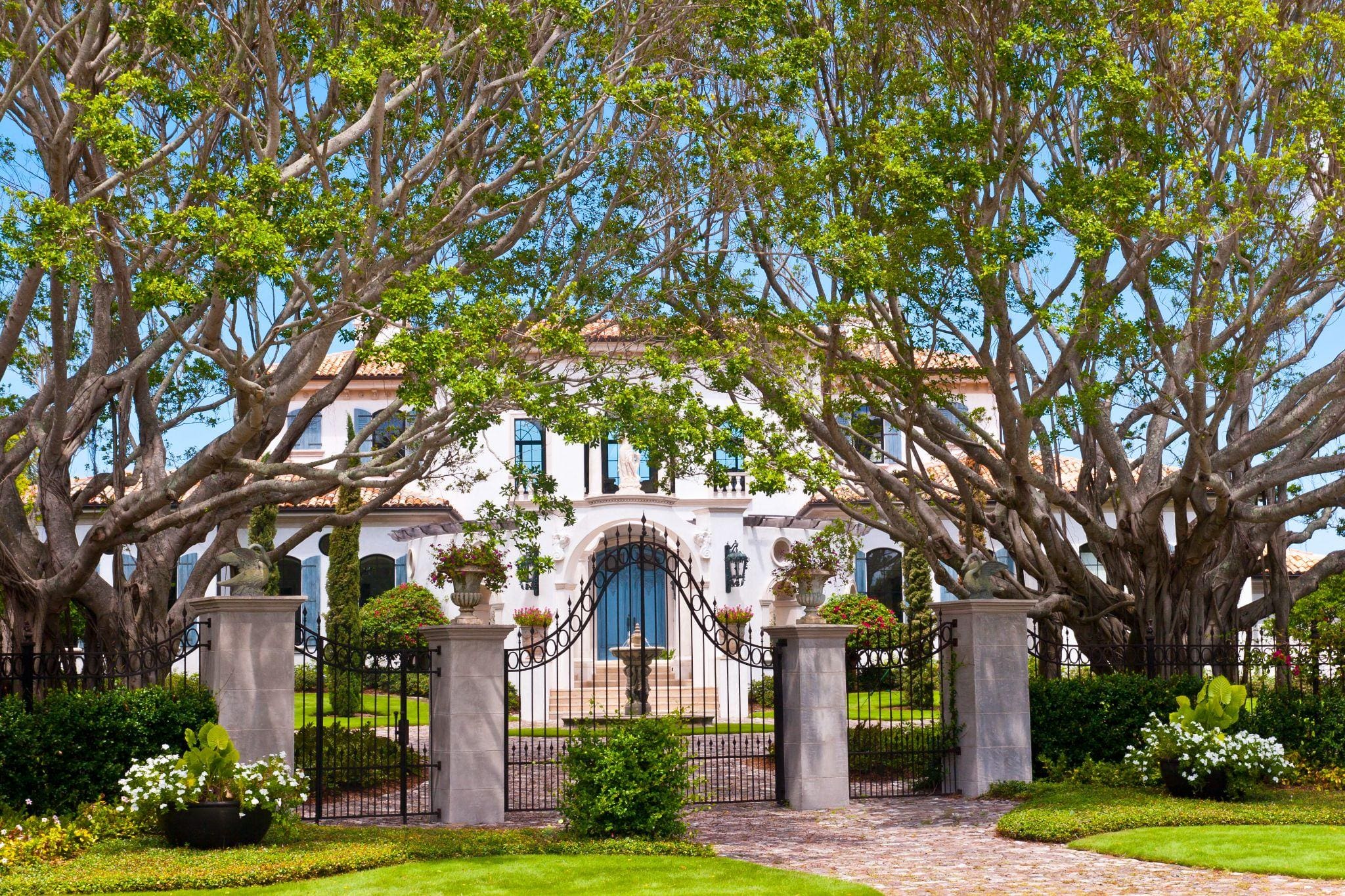 A stunning entrance to a luxurious home, framed by large trees and a wrought-iron gate, showcasing a beautifully landscaped garden.