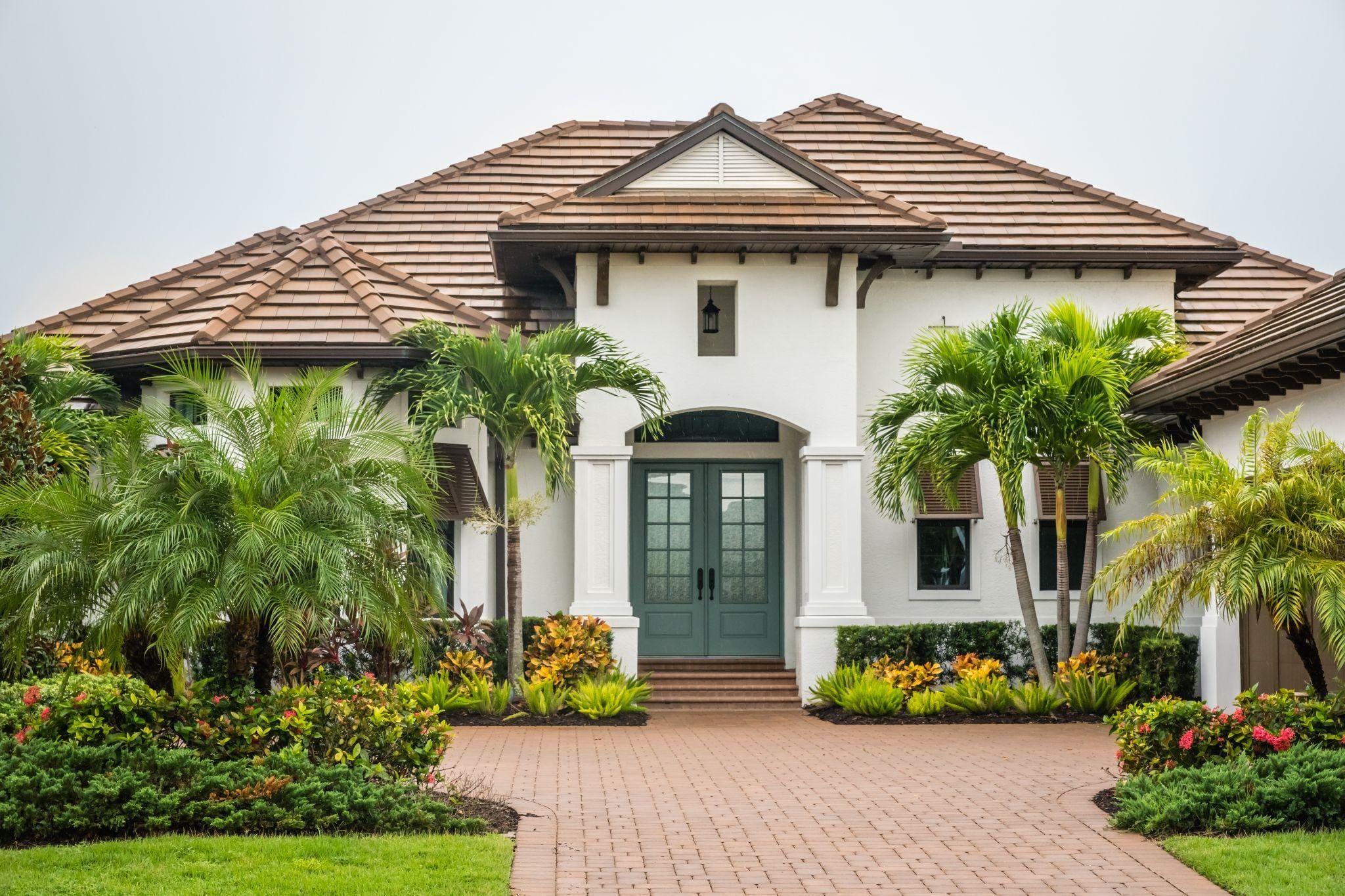 An elegant two-story house with a well-kept lawn, surrounded by lush greenery and a brick pathway leading up to the entrance.