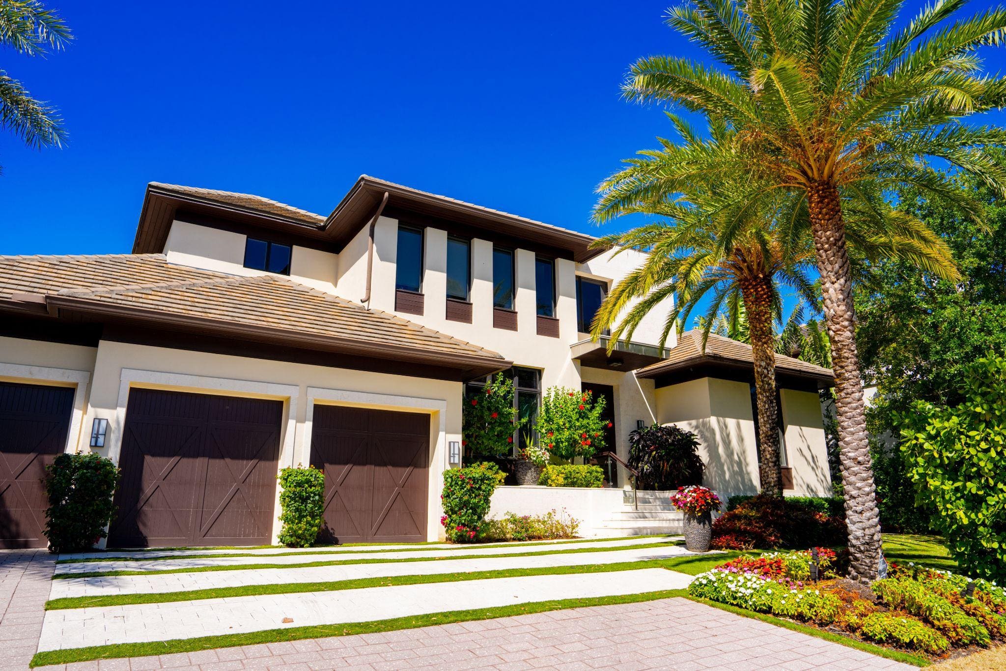 A modern two-story house with a stylish facade, featuring large windows, a well-maintained garden, and palm trees, all under a clear blue sky. The driveway is lined with decorative stones and vibrant flowers.