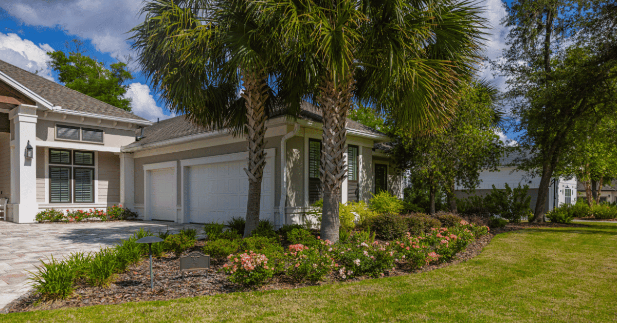 Exterior view of a single-story home on a sunny day, featuring light-colored siding, white trim, and a paved driveway leading to a white garage. The lush front yard is landscaped with prominent palm trees, green shrubs, and pink flowering bushes bordering a green lawn.