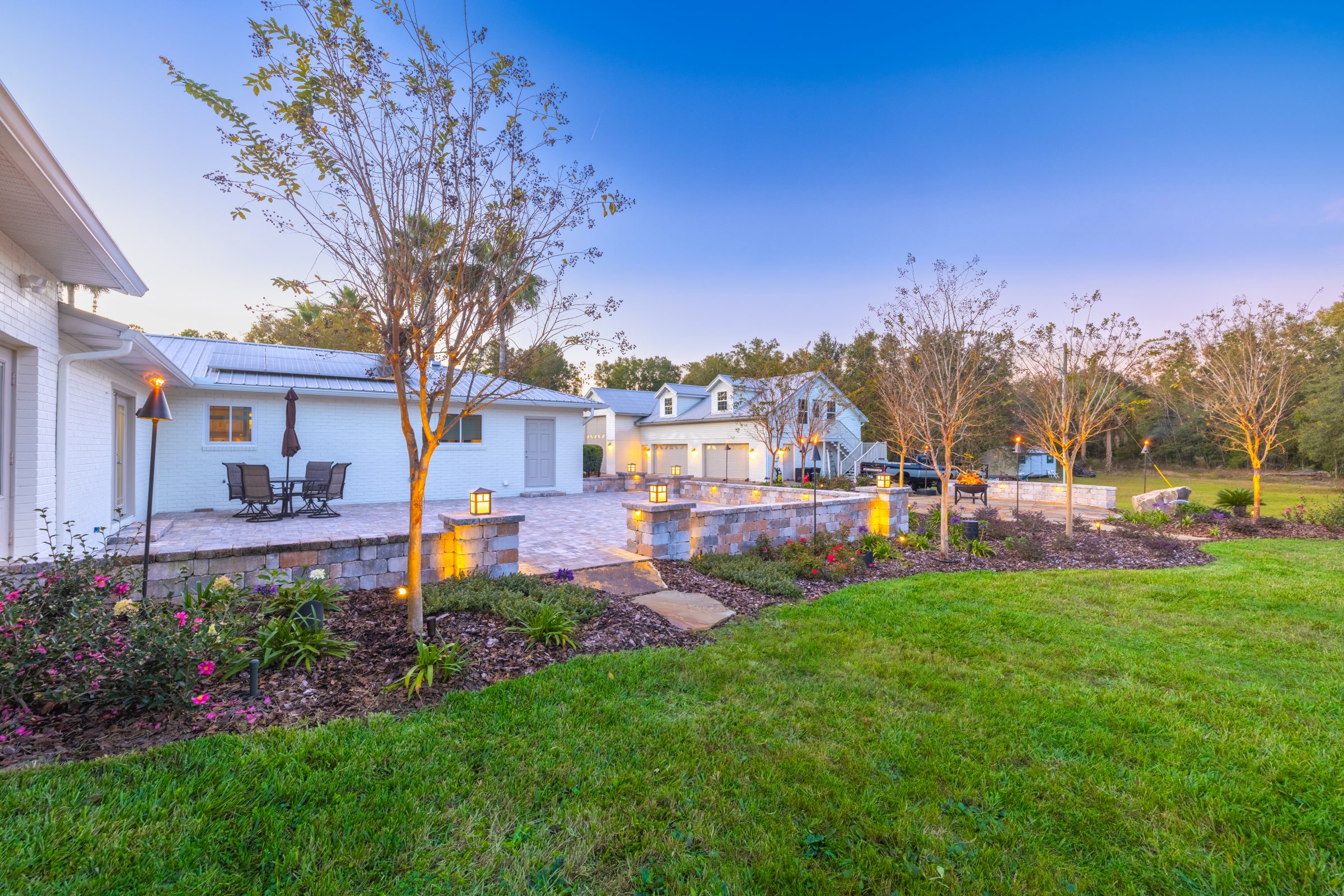 Illuminated landscaped patio beside white brick home at dusk