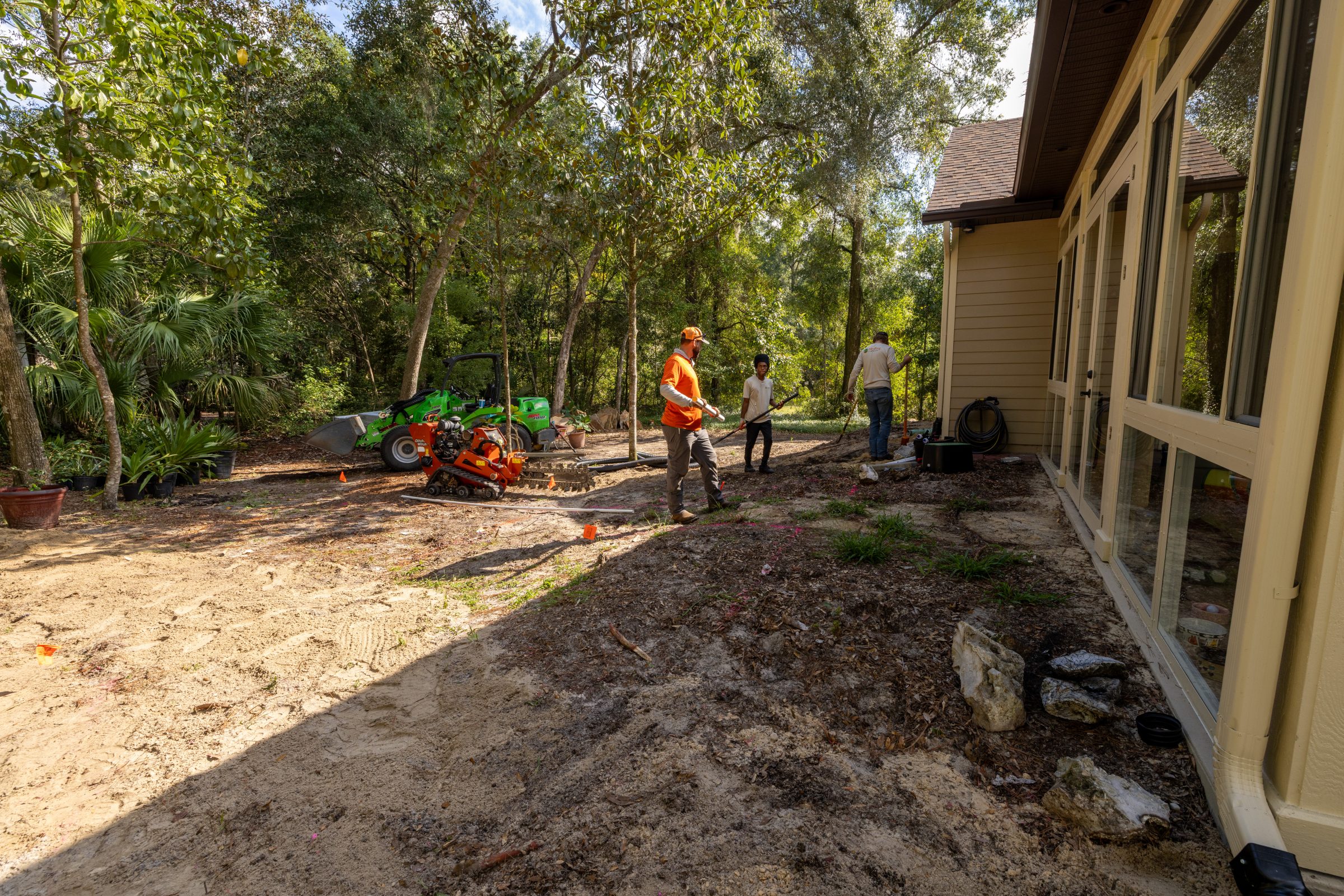 Landscaping crew prepares wooded yard beside a beige house.