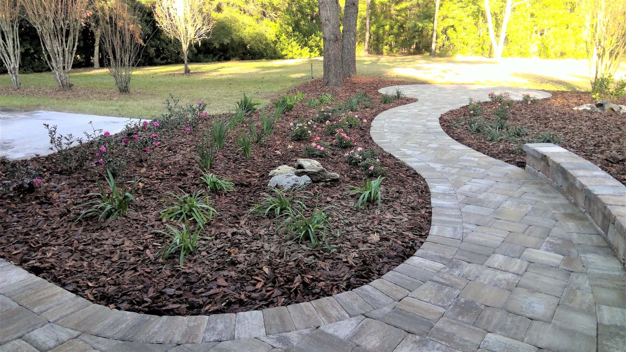 Curved paver stone walkway bordering a freshly mulched garden bed with young plants and flowers, leading toward a grassy yard.