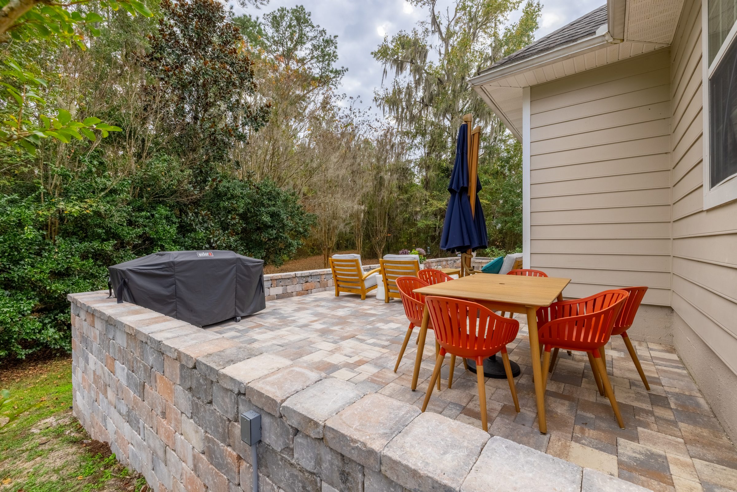 Paved backyard patio with dining table, grill, and wooded view