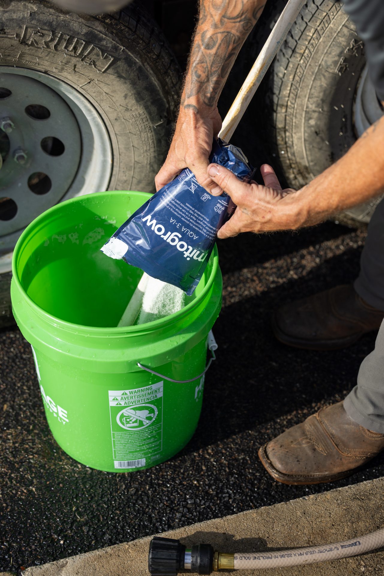 Worker pours cleaning powder into green bucket beside truck tires.